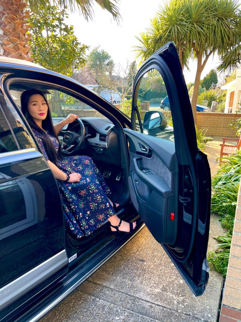 Lei Hang sitting in a black Audi Q7 S-line, wearing a navy blue floral dress with black sandal heels. Will she swap her car for a £3000 per year mobility credit to become more eco-friendly? 