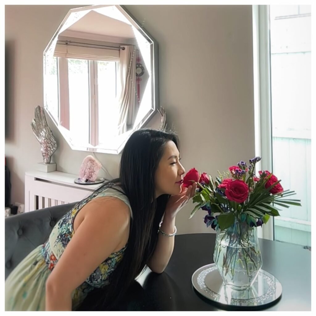 Lei Hang aka the thrifty island girl as seen on channel 5 tv show bargain brits on benefits is smelling a rose from a bunch of flowers on her black dining table for a virtual facetime photoshoot